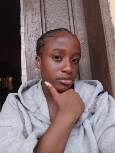 A young Black woman poses for a selfie. She is wearing a gray hooded sweatshirt. Her hand is resting on her chin in a pensive gesture. She has braids. Behind her, there is a partially open doorway leading into a dark space and a textured metal door. There is no information to confirm the location is in Umuahia, Nigeria.