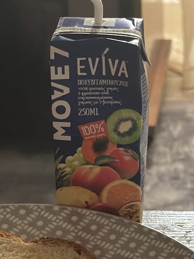 A vertically oriented, close-up shot showcases a carton of "MOVE7 EVIVA" fruit juice. The carton is predominantly blue and features various fruits depicted on its surface, including grapes, apples, a kiwi, and oranges. Text on the carton indicates "100% φυσικές χυμές" (100% natural juices) and "9 φρούτων από συμπυκνωμένους χυμούς με 7 βιταμίνες" (9 fruits from concentrated juices with 7 vitamins), along with the volume "250ML." A red label with "100%" is also visible. The carton is positioned on a grey and white patterned surface, with a portion of what appears to be bread or toast in the foreground. The background is blurred, suggesting a domestic or indoor setting, and is not specific enough to identify the location in Dromolaxia - Meneou Municipality, Cyprus.