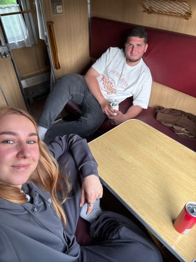 The image captures two individuals seated at a table in what appears to be the interior of a train or similar transportation vehicle. The man is seated on a burgundy-colored seat, wearing a white t-shirt with a print that includes the word "PICNICLOVER," and dark grey jeans. He is holding what appears to be a white cylindrical object. The woman, closer to the camera, is wearing a dark grey hooded sweatshirt and the angle indicates she may be taking the picture. A can of soda sits on the table. The environment suggests travel, possibly within the area of Радашковіцкі сельскі Савет, Belarus. A brown jacket is lying on the seat near the table.