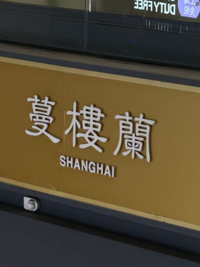 A close-up, angled, medium shot shows white 3D Chinese characters and the word "SHANGHAI" on a textured gold background. The Chinese characters read "蔓樓蘭". The characters are raised from the gold surface. Below the Chinese characters, the English word "SHANGHAI" is spelled out in all capital letters, also in white and raised from the gold surface. The gold surface is bordered by a dark gray band on the bottom. A small, circular, silver security camera is visible on the dark gray band in the lower left corner of the frame. In the background, a portion of a dark reflective surface with some blurred white text is visible at the top. The overall scene appears to be signage, likely in a public space in Shanghai, China, potentially at an airport or a commercial establishment in the Pudong district given the context of Shanghai.