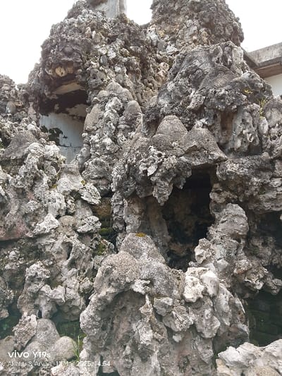 The image shows a close-up of a rock formation in Cirebon, Indonesia, located at the coordinates -6.736 latitude and 108.544 longitude. The formation consists of irregularly shaped, grey-colored rocks with a textured surface. There are small openings in the rock formation, suggesting a possible cave or grotto. The rock surface is covered in small patches of green moss and other plant life. The rocks are arranged in an irregular pattern, and there are no people or animals present in the image. The background shows the sky, indicating an outdoor setting.