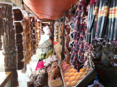 This image depicts a bustling market stall, likely in Pantegakibale, Indonesia, during daytime. An orange tarp canopy stretches overhead, providing shade to the diverse array of goods on display.
The stall is laden with various products, many individually packaged in clear plastic bags and hung from ropes or hooks. On the left, rows of dark, flat, irregularly shaped items, possibly dried meat jerky or fruit leather, are prominently featured, with some bags bearing small yellow labels that read "Cara_Yha" and include a logo. Further along, other packaged snacks, some labeled "GURIH NIKMAT PARADE," are also suspended.
In the mid-ground, a vibrant assortment of items is arranged on tiered shelves. This includes piles of green, spherical fruits (possibly unripe mangoes or limes), along with numerous white, soft, oval-shaped items that could be a type of sweet or snack. Bright pink, fluffy materials are also visible, alongside various packaged baked goods or local delicacies. In the background, large white sacks are stacked, suggesting bulk storage.
To the right, more hanging merchandise dominates the foreground. Bundles of long, slender, dark sticks, possibly incense or traditional sweets, are neatly arranged, tied with red and yellow bands. Nearby, distinctive clusters of smaller, white, tightly wrapped packets, secured with red ties, hang alongside. Beneath these, a large display board prominently features an image of ripe yellow mangoes, with handwritten text partially visible, including "BL-1167-AN" and "UMAR," as well as what appears to be "02-23." A partial view of a person's arm and shoulder is visible on the far right, hinting at activity within the market. The scene conveys a lively market environment offering a wide variety of local produce and packaged goods.