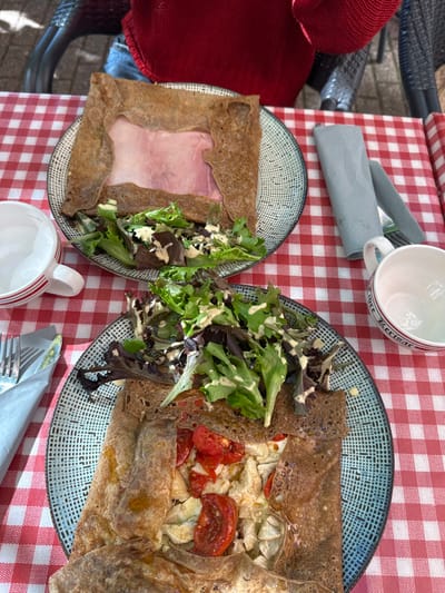 Two savory crepes (galettes) with accompanying side salads are displayed on a red and white checkered tablecloth in an outdoor dining setting in Orléans, France. The upper plate presents a square-folded buckwheat crepe containing a prominent slice of ham, beside which lies a portion of mixed green salad with a creamy dressing. The lower plate features a similar buckwheat crepe filled with chopped white meat, halved cherry tomatoes, and possibly artichoke hearts, also accompanied by a mixed green salad with dressing. Both plates are dark blue and white speckled ceramic. An empty white ceramic cup with red horizontal stripes rests to the left of the upper plate. Another empty white ceramic cup, with a red rim partially inscribed with "fore Kerisac," is situated to the right of the lower plate. A light blue-gray folded paper napkin, concealing cutlery, is placed on the upper right of the table, and another similar napkin with visible cutlery is on the lower left. A person wearing a red knitted garment and blue jeans is partially visible in the background, seated on a dark, woven-style chair behind the upper plate. The arrangement indicates a meal prepared for at least two individuals.