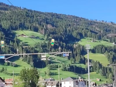 The image shows a view of a green mountainside in Innichen - San Candido, Italy, with a clear blue sky. The mountain is covered with dense evergreen trees and dotted with small buildings, suggesting a rural or semi-rural setting. In the foreground, there are signs and structures that appear to be related to transportation infrastructure, possibly for a cable car or ski lift system, given the presence of a tall tower with a gondola. The lower part of the image reveals a few houses with red roofs, typical of alpine architecture. The scene is illuminated by bright sunlight, casting shadows on the grassy slopes.