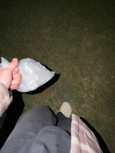 The image shows a person holding a clear plastic bag filled with an opaque substance. The person is wearing dark jeans, a light-colored shoe, and a plaid jacket. The background appears to be a dark grassy area. The lighting suggests it is nighttime.