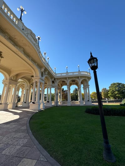 The image shows a long, white colonnade structure under a bright blue sky in Tigre, Argentina. The architecture is ornate with arches and decorative elements. Street lamps are affixed to the top of the structure and line the walkway in front of it. The ground is paved with rectangular stones. Green grass and shrubbery border the walkway. A taller black street lamp is positioned in the foreground against the blue sky. Trees are visible in the background.