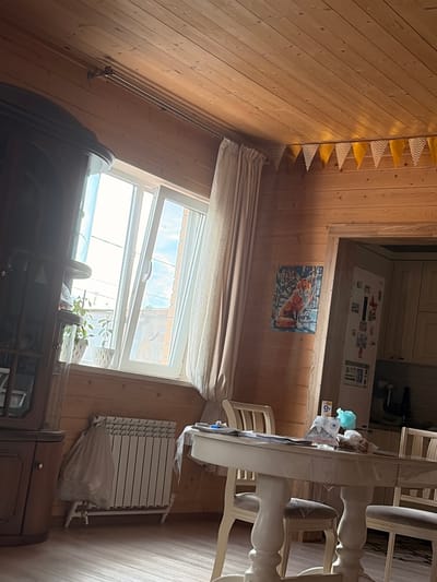 The image depicts the interior of a room with wooden paneled walls and ceiling, likely a residential space in Leninogorsk, Russia. A large window with white frames is visible on the left, letting in bright daylight and showing a glimpse of the sky and possibly outdoor structures. In front of the window, there is a white radiator covered with a translucent plastic bag. To the left of the window, a tall, dark wooden cabinet or hutch is partially visible.
In the center of the room, a round white dining table with carved legs is set with several chairs. The table surface has some items on it, including what appears to be a plastic tablecloth, papers, and a few small objects. To the right of the table, a doorway leads to another area, likely a kitchen, where a refrigerator with magnets on its door and some cabinetry are visible.
On the wooden wall to the right of the window, a framed picture of a fox sitting in a snowy landscape is hung. Further up the wall, near the ceiling, a string of triangular yellow and white pennant banners is displayed. The floor is made of light-colored wood planks. The overall impression is of a lived-in, rustic interior. There are no people visible in the image.