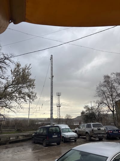 The image depicts an outdoor scene in Cherven Bryag, Bulgaria, under an overcast, grey sky. The perspective is from beneath a yellow corrugated metal awning or overhang, from which water droplets are visible.

The foreground consists of a wet, unpaved or gravel parking area. Parked vehicles include, from left to right: a dark blue Peugeot Partner minivan (license plate CB 4121 AO), a white Renault Kangoo or similar small van (license plate EH 0142 BB), a silver/grey Nissan Navara pickup truck with a bed cap, appearing modified with lifted suspension and off-road tires, splattered with mud (license plate EH 9000 KT), and a dark blue Volkswagen Passat sedan (license plate EH 6178 KC). The front of a fifth silver car is partially visible in the bottom right corner.

Behind the vehicles, railway tracks are visible, accompanied by an overhead electrical catenary system supported by metal poles. Adjacent to the tracks, the ground is covered with bare deciduous trees and brush.

Prominently featured in the mid-ground is a tall, slender metal lattice mast or communication tower, equipped with multiple guy wires and a light fixture. A smaller, similar lattice tower is visible further in the background to the right of the main tower. On the left, a deciduous tree displays sparse, light-colored blossoms or early foliage, while trees on the right are bare.

In the distant background, rolling hills are partially obscured by atmospheric haze or low clouds. No individuals are visible in the scene, and no actions or interactions are depicted.