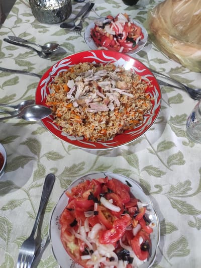 A top-down, medium shot captures a meal laid out on a table. The setting appears to be a dining room in Mingchinar, Uzbekistan, indicated by the traditional patterns on the tablecloth and serving ware. The tablecloth is cream-colored with a repeating pattern of green leaves and stems.
The primary subject is a large, round, red plate filled with pilaf, a rice dish cooked with meat, carrots, and spices. Shredded chicken is piled on top of the pilaf. To the right of the pilaf, a white plate holds a tomato and onion salad, garnished with dark herbs. Another similar salad bowl is visible in the bottom right corner of the image.
Several pieces of cutlery, including spoons and forks, are scattered around the plates, suggesting a meal in progress or ready to be served. A glass jug with intricate metalwork is visible in the background, and a portion of flatbread wrapped in plastic is to the upper right. The lighting suggests it might be evening or indoors. The overall impression is of a home-cooked meal, possibly a family gathering or a shared meal with guests. No visible text is present.