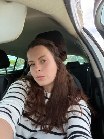 A young woman is captured in a full-frame portrait, looking directly at the camera with a neutral expression. She has long, wavy brown hair and is wearing a white sweater with black horizontal stripes. The photograph appears to be a selfie taken from inside a car, likely from the passenger or back seat, given the upward angle.
The car's interior is visible, featuring cream-colored headliner and seat fabrics, along with black trim and dark-colored seats. Through the car window behind the woman, an outdoor scene is discernible under what appears to be an overcast daytime sky.
Prominent in the background is a red sign with white lettering, clearly showing the word "IKEA." Below "IKEA," additional partial text suggests the name of the location, consistent with Kaiserslautern, Germany, as provided in the context. A blue and white "P" parking sign is also visible, indicating the car is situated in a parking area. Green foliage from trees and parts of a building structure can be seen outside the window. The overall lighting suggests it is daytime.
