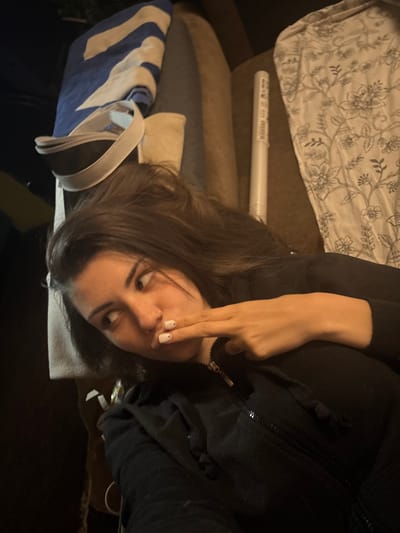 The image portrays an indoor scene, likely a bedroom or living room, characterized by dim lighting, suggesting evening or nighttime. The central subject is a young woman with dark brown hair, seen reclining or lying down on a piece of furniture, possibly a couch or bed. She is wearing a black long-sleeved top or hoodie, which features a visible zipper and drawstring details. Her right hand is positioned near her mouth or chin, with fingers slightly curled, and her fingernails are painted a light color, possibly white or pale pink, each adorned with a small, darker dot. She looks towards the right side of the frame with a neutral to subtly pensive expression.
In the upper part of the image, behind her head, is a light-colored sheet or duvet cover patterned with a grey or black floral and botanical design. Below this, a long, white cylindrical object, possibly a tube for a poster or artwork, is visible. It bears small, indistinct text and symbols on its surface, which are not clearly legible. Further to the left and foreground, a blue and white striped bag or fabric item with prominent white handles rests on dark-colored furniture. The overall composition suggests a relaxed, personal moment within a residential setting in Belopoptsi, Bulgaria, though no specific geographical identifiers are present within the visual content. The original image appears rotated 90 degrees counter-clockwise.