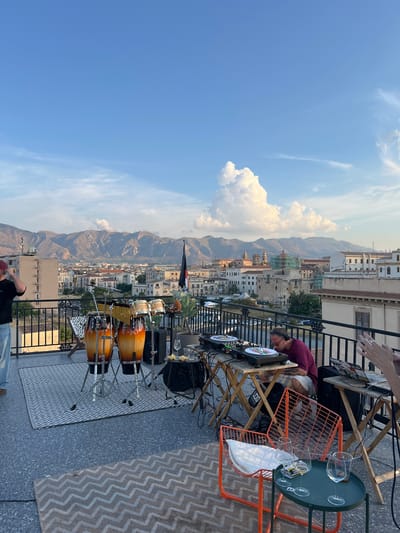 This media file captures a rooftop scene with a DJ setup and percussion instruments against a backdrop of Palermo, Italy. Two people are visible: one man, wearing a maroon shirt and headphones, appears to be DJing, interacting with turntables and mixers. His posture suggests focus. Another person, wearing a red cap, is partially visible in the lower frame, appearing to be relaxed.
The scene is set on a tiled rooftop with a railing overlooking a densely built cityscape that extends to mountainous terrain. The architecture of the buildings suggests an Italian location, consistent with the provided context of Palermo. Percussion instruments, specifically conga drums, are arranged on stands, ready for use. A small side table holds wine glasses and what appear to be cigarette packs.
The time of day appears to be late afternoon or early evening, indicated by the warm sunlight casting long shadows and the color of the sky. The sky is a clear blue with scattered white clouds, suggesting fair weather.
Notable details include the intricate patterns on the rugs placed on the rooftop floor and the detail of the orange metal chair. The overall mood seems to be relaxed and perhaps anticipatory of a musical performance. No visible text is clearly legible in the media file.