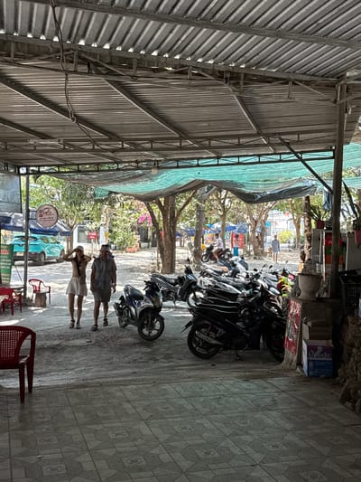 The image shows a street scene in Diên Khánh, Vietnam. The view is taken from under a corrugated metal roof structure, partially covered with a green tarp. Numerous motorcycles are parked haphazardly on the right side of the frame. Two people, a man and a woman, are walking along the street. In the distance, trees line the street, and various commercial signs are visible. Red plastic chairs are present near the bottom of the frame, suggesting a small outdoor dining area. The overall scene conveys a typical streetscape in a Southeast Asian town.