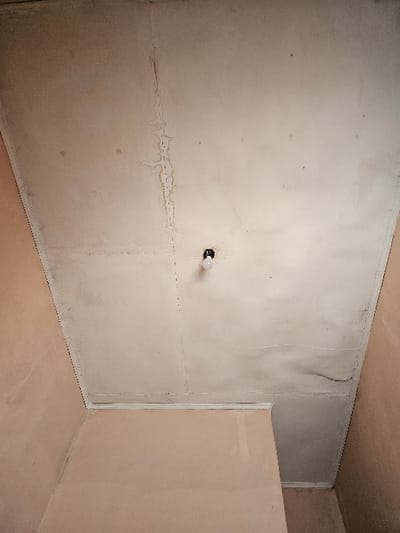 The image displays an interior view of a room in Muka, Indonesia, looking upwards towards the ceiling and upper walls. The light-colored ceiling, appearing to be constructed from panels, shows significant signs of water damage, most notably a prominent dark brown, winding stain running vertically down the left half, with several smaller branched stains. A single, bare lightbulb with an exposed filament is screwed into a black fixture, suspended from the ceiling in the lower right portion of the frame. Visible seams demarcate the ceiling panels. The upper sections of the walls are uniformly painted in a light peach or salmon tone, meeting the ceiling with a simple, light-colored trim. A section of an adjacent or partition wall, also peach/salmon, is visible in the lower right, forming an interior corner. There are no people present.