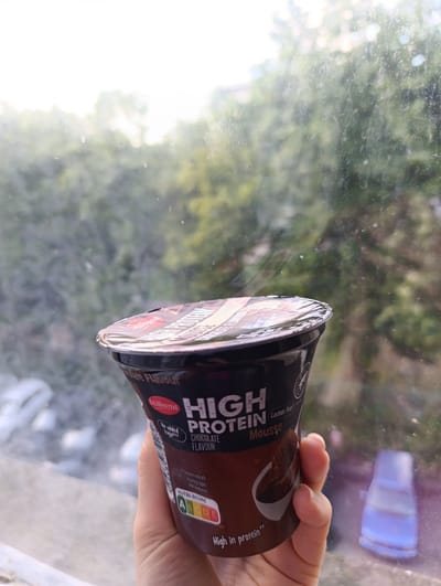 A hand holds a Milbona High Protein Chocolate Flavour Mousse container in front of a window. The container is dark brown with text and a logo visible. The background shows blurred greenery and a white sky, suggesting an outdoor scene viewed from indoors. There are no other people or discernible landmarks from Razgrad, Bulgaria present in the image. The lighting suggests it is daytime.