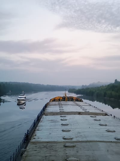 This image depicts a tranquil scene on a waterway, likely the river at Volokov Most, Russia. In the foreground, the deck of a large barge, characterized by its flat surface and numerous circular hatches, stretches towards the viewer. A blue railing runs along the side of the barge, delineating its edge. To the left, on the calm water, a sleek white yacht with a black stripe is navigating, leaving a visible wake. The yacht is positioned in the middle ground, moving away from the perspective of the viewer. The surrounding environment is characterized by lush green trees lining the banks of the waterway, creating a dense, forested landscape. The sky above is overcast with a soft, diffused light, suggesting either early morning or late evening. The overall atmosphere is serene and quiet.