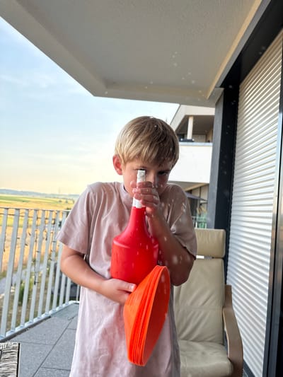 A young boy stands on a balcony, holding a red spray bottle in front of his face and a stack of orange cones in his left arm. He is wearing a light brown t-shirt. The background shows a landscape with fields under a partly cloudy sky. To the right of the boy, a cream-colored chair is visible in front of a closed, ribbed shutter. Water droplets are dispersed throughout the air around the boy. The balcony railing is white and made of vertical bars. The scene appears to be taking place in Bönnigheim, Germany, on a balcony overlooking a rural area.