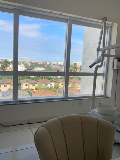 The image depicts an interior scene looking out through a large, multi-pane window from an elevated position. The interior foreground features the light-beige, ribbed upholstered backrest of a chair, likely a patient's chair. The floor is composed of white tiles, with a black cable running along the base of the wall. To the right, a white dental unit is partially visible, distinguished by a ceramic spittoon bowl and an articulated grey arm extending vertically from its base, characteristic of an examination light or X-ray apparatus. A white electrical receptacle is affixed to the wall below a dark granite windowsill.
The exterior view through the window reveals a clear blue sky with scattered white clouds. In the middle ground, a residential area is visible, consisting of multiple single-story homes with light-colored facades and reddish-brown tiled roofs. Several of these roofs are equipped with photovoltaic solar panels, and some feature white satellite dishes. Green trees and dense vegetation, including palm trees on the left, are interspersed among the residences. The distant background displays a denser urban skyline, composed of numerous multi-story buildings and high-rises. A tall, white building with a grid-patterned exterior is prominent immediately to the right of the window frame.
This scene is located in Lauro de Freitas, Brazil. The specific location is consistent with an upper-floor view from a commercial establishment, overlooking a mix of suburban residences and a developing urban core, indicative of areas such as **Vilas do Atlântico** or the **central commercial districts** of Lauro de Freitas. No individuals are present in the image.