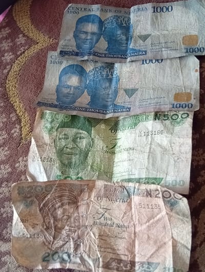 The image displays four worn and creased Nigerian Naira banknotes arranged on a brown and red patterned textile surface, likely a rug or carpet, in Zango, Nigeria. From top to bottom, the visible currency includes two 1000 Naira notes (one dated 2023, the other 2022, both featuring portraits of Aliyu Mai-Bornu and Clement Isong), one 500 Naira note (dated 2025, featuring Dr. Nnamdi Azikiwe), and one 200 Naira note (dated 2021, featuring Sir Ahmadu Bello). The banknotes show signs of heavy use, characterized by numerous folds and creases. No individuals or direct actions are depicted; the scene focuses solely on the currency and its immediate environment.