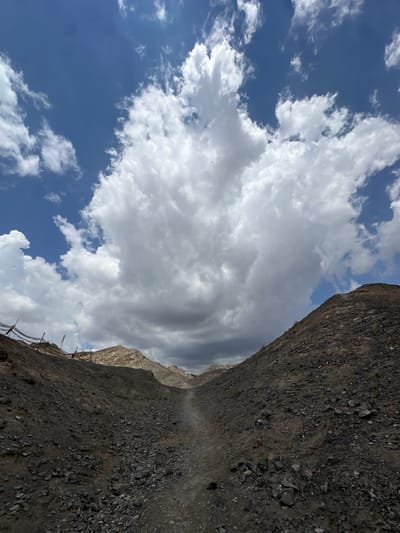The image depicts a barren, rocky landscape with a narrow dirt path winding through the center, leading towards distant mountains under a dramatic sky filled with large, fluffy white clouds and patches of blue. The terrain is covered in small, dark grey rocks and sparse vegetation. On the left side of the path, a low fence with colorful prayer flags is visible, suggesting a cultural or religious significance to the location. The mountains in the background are arid and appear to be made of layered rock formations. The overall scene conveys a sense of rugged, natural beauty and solitude. The image's composition and subject matter suggest it could be a hiking trail or a scenic route in a mountainous or desert region. This type of landscape is characteristic of areas near Jiayuguan, China, particularly in its surrounding natural parks or historical sites, such as those near the Great Wall of China.