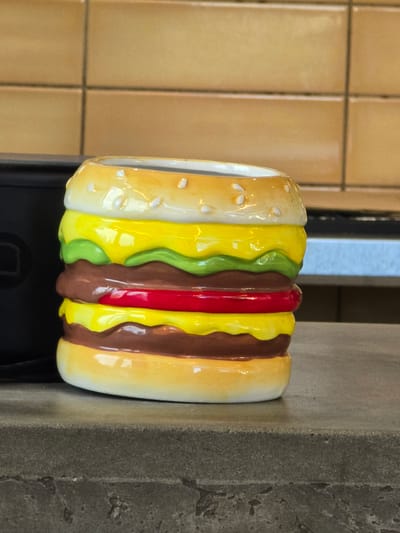 A ceramic hamburger-shaped mug sits on a gray, textured countertop. The mug is layered with yellow bun halves on the top and bottom, with white sesame seed details on the top bun. The interior of the mug is white. Between the bun halves are layers representing cheese (yellow), lettuce (green), a beef patty (brown), and a tomato slice (red). The background consists of light brown tiled walls and a blurred dark object to the left. The scene appears to be indoors, possibly in a kitchen or restaurant setting. The location is specified as Duluth, United States of America.