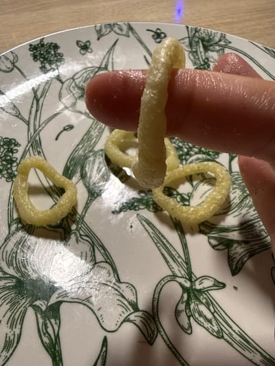 A person's index finger, coated in a glistening substance, is inserted into a yellow, ring-shaped snack food. The snack is textured with spiral ridges and appears somewhat brittle. The finger is positioned against a white plate decorated with green line drawings of botanical motifs, possibly irises and other flowers, and small butterfly-like insects. Several other similar snack rings are scattered across the plate. The background shows a wooden surface. The scene is illuminated by artificial light, which creates a slight blue reflection on the plate. There is no information to suggest this scene is located in Cheboksary, Russia.