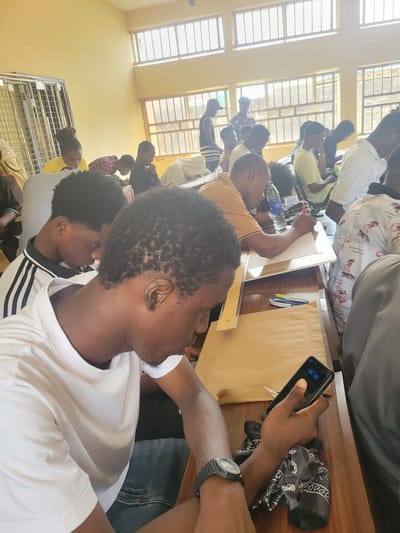 This media file shows a group of young people gathered around tables in what appears to be a classroom or workshop setting. The individuals are predominantly male and are of African descent. Many are wearing casual clothing such as t-shirts and jeans.
The scene depicts a collaborative activity or learning session. Some individuals are leaning over tables, looking at papers or devices. One person in the foreground is holding a smartphone, suggesting technology is in use. Another person is seen working with a ruler and a piece of paper, possibly engaged in drafting or drawing. A bottle of water is visible on one of the tables.
The environment is brightly lit, suggesting it is daytime. The room has yellow walls and features large windows with gridded panes, typical of educational institutions. A ceiling fan is partially visible, indicating the need for cooling. The overall atmosphere seems focused and engaged, with individuals concentrating on their tasks. The clothing styles and the overall setting are consistent with a location in Ukukwa, Nigeria. No discernible text is clearly visible in the image, apart from what might be on the phone screen.