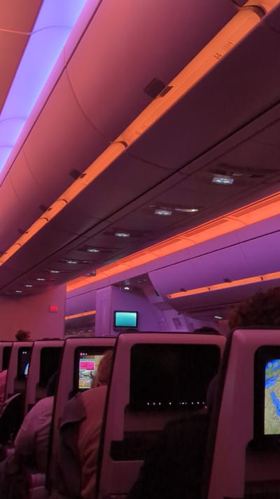 The image shows the interior of an airplane cabin at night, with purple and orange lighting. The view is from a passenger's perspective, looking upwards and towards the front of the cabin. Rows of seats with individual entertainment screens are visible. The overhead compartments and lighting fixtures are illuminated. Several passengers are seated, with some looking at their screens. A flight attendant call button is visible on the wall. The scene appears to be in an airplane cabin, likely during a flight. The location of Unknown, United Arab Emirates is irrelevant to the visual content of the image.