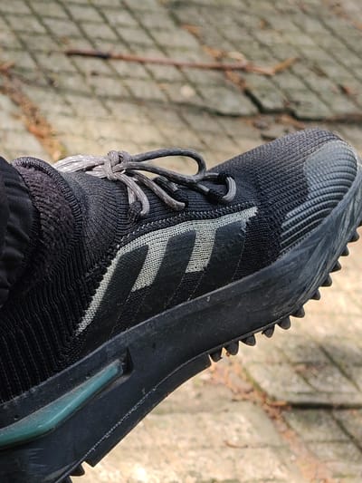 An individual's left foot, encased in a black athletic shoe, is captured in a close-up, angled view within an outdoor environment in Tsarev Brod, Bulgaria. The shoe features a black, knitted fabric upper with a seamless, sock-like ankle collar. Grey shoelaces are tied with a knot on the dorsal aspect of the foot. Three parallel, diagonal grey stripes, consistent with Adidas branding, are visible on the lateral side of the shoe's upper. The black toe box is reinforced with a horizontally ribbed texture. The black midsole incorporates a distinct teal-colored rectangular insert on its medial side. The black outsole is equipped with a pattern of prominent, pointed lugs, indicating suitability for varied terrain. The shoe exhibits signs of moderate wear and dust accumulation. The blurred background consists of light-colored, irregular paving stones or concrete slabs on the ground, with scattered natural debris such as dried leaves. The scene is illuminated by natural daylight. No other individuals or specific activities are depicted.
