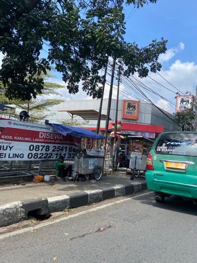 The scene is a street in Cimahi, Indonesia, located at coordinates -6.886 latitude and 107.554 longitude. A green car is parked on the side of the road. There is a sidewalk and a curb separating the road and sidewalk. Behind the curb is a fence with a sign that says "Disewa" and a phone number. There are two people sitting on the sidewalk next to a food cart. The cart has a variety of snacks for sale. In the background, there is a supermarket named "Super Indo" on the corner of the street. There are trees and power lines overhead. The sky is clear and blue.