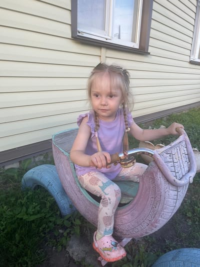 A young Caucasian girl with blonde hair in two braids is sitting on a toy vehicle made from painted tires. She is wearing a light purple t-shirt and patterned leggings. Her right hand is on the handlebars of the toy, which is decorated with a light blue and pink color scheme. The toy is situated on a grassy area with some visible rocks and a concrete border. In the background, a light-colored, horizontal-sided building with windows is visible. The image appears to be taken outdoors in daylight.