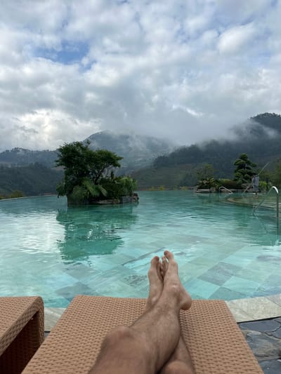 The image depicts an outdoor infinity pool scene located in Púng Luông, Vietnam, under a cloudy sky. In the immediate foreground, a person's bare, hairy feet are crossed at the ankles and resting on a light brown, woven lounge chair, indicating relaxation. The pool, tiled with light turquoise and grey squares, occupies the midground, its surface showing gentle ripples.
Within the pool, two decorative islands are visible. The larger island on the left features a dense cluster of green trees and broad-leafed plants, reflecting in the water. On the right side of the pool, a smaller island hosts a meticulously pruned bonsai-style tree and additional greenery. Further right, a male figure wearing a grey hooded top and shorts stands in the pool, holding a long pole, suggesting pool maintenance activity. Stainless steel handrails are visible on the pool's edge in the far right.
The background is dominated by a expansive mountainous landscape covered in dense green foliage. Low-hanging clouds and mist partially obscure the mountain peaks, creating a hazy atmosphere. Terraced hillsides, characteristic of agricultural cultivation, are discernible on the left slopes. The sky above is largely overcast with white and grey clouds, though a small patch of blue sky is visible towards the top center.