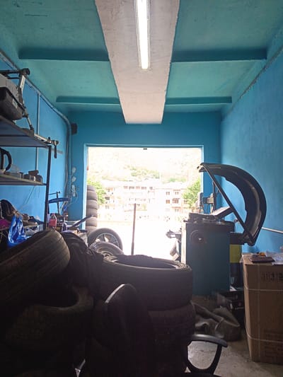 This image depicts the interior of a tire repair shop in Kapan, Armenia. The scene is brightly lit by a fluorescent light fixture on the ceiling and natural light streaming in from the open doorway. The walls and ceiling are painted a vibrant turquoise blue.

The foreground is dominated by a pile of used tires, some stacked high, others lying in disarray. To the right, a tire changing machine with a large, dark curved arm is prominent. Next to the machine, a cardboard box is stacked on top of another, with some paper on top.

In the background, the open doorway reveals a view of a town nestled against a hillside. Buildings with light-colored facades and trees are visible under a bright, overcast sky, suggesting it is daytime.

On a shelf on the left side of the image, various items are stored, including what appears to be a tire pressure gauge, bottles, and other tools. A blue plastic bag is also visible amongst the items. A red bottle of liquid and a metal stand with the word "ROSSVIK" are also present.

The overall impression is a working garage, likely a small business. The bright colors and natural light create a somewhat cheerful, though busy, atmosphere. There is no visible text that is clearly legible, except for the "ROSSVIK" label.
