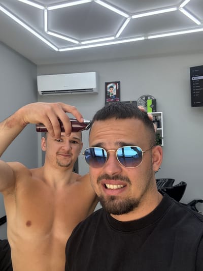 An indoor mirror selfie shows a man smiling broadly into the camera. He has short dark hair, a beard, and wears gold-framed sunglasses with blue reflective lenses (partially revealing "Ray-Ban" on the temple) and a black t-shirt. His reflection, seen in the lower part of the image, shows him shirtless, holding a dark red hair clipper near his head, also with a slight smile.
The background features a modern, clean room. On the left wall, hexagonal LED light fixtures provide bright illumination. A white "GREE" air conditioner unit is mounted on the wall. To the right, a flat-screen TV displays a menu with options including "Sign in," "Search," "Gaming," "Subscriptions," and "Settings." Other visible decor includes a framed picture and a circular wall ornament. The man's cheerful expression suggests a positive mood. The scene is set indoors, possibly in Simitli, Bulgaria.