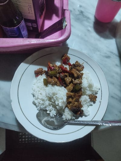 A white plate with a portion of white rice and a stir fry with meat, vegetables, and sauce. A stainless steel spoon is lying next to the plate, with the handle extending off the right side of the image. The plate sits on a white marble table with a partially visible pink plastic container in the upper left corner of the frame. The location is Mojokerto, Indonesia (latitude: -7.459, longitude: 112.447).