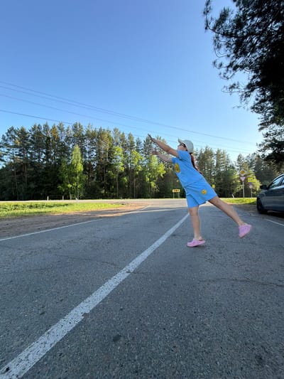 A woman in a light blue shorts and t-shirt set with smiley face patches is lying horizontally on a paved road surface with a white line divider. She has a light-colored baseball cap and pink shoes. Her arms are outstretched. In the background, there are tall green trees and a clear blue sky. In the upper right corner, a portion of a dark-colored vehicle is visible. Roadside infrastructure, including road signs, can also be seen. This scene is located in the city of Foki, Russia.