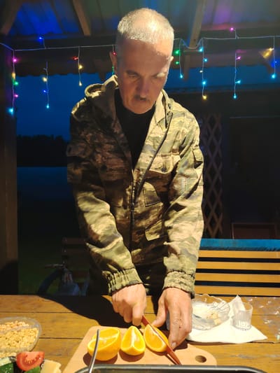 An older man with gray hair and a mustache is cutting oranges on a wooden table. He is wearing a camouflage jacket and a dark shirt. In front of him on the table are a bowl of peanuts, a tomato slice, a piece of cheese, and a cutting board with two orange slices. There is also a small bowl with some food residue and a shot glass on a white napkin. Behind him, colorful string lights are visible. The scene appears to be outdoors, possibly on a porch or patio, with a dark background suggesting it is evening. The setting is the city of Novaya Prismara, Russia.