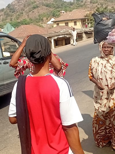 A woman carries a bag on her head while standing next to an older woman in patterned clothing, who also carries a bag on her head. Another woman in white shorts and a red top stands with her back to the camera. They appear to be on a roadside with buildings and a hillside in the background. The photo is taken at an angle.