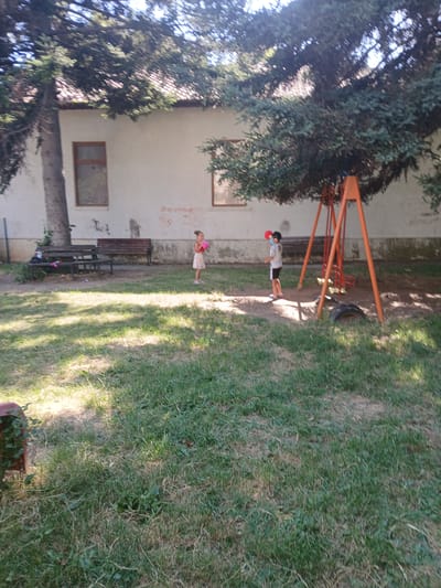 In Obrochishte, Bulgaria, a sunny daytime scene unfolds in a park or playground area. Two children are visible, one standing by an orange swing set, and another, a girl, holding a pink ball. The setting includes a grassy lawn, a building with windows, and a tree providing shade. The overall atmosphere appears to be relaxed and playful. There is no visible text in the image.