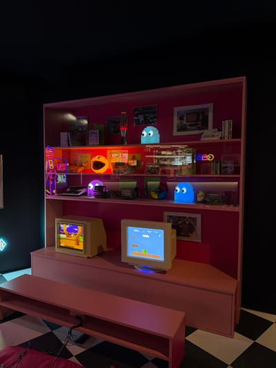 This scene, located in Lausanne, Switzerland, depicts a retro video game display. A prominent, freestanding magenta-pink shelving unit is positioned against a dark background.

On the uppermost shelf, from left to right, are multiple framed photographs, a red lava lamp, a light blue pixelated Pac-Man ghost lamp, and a stack of books or media.

The middle shelf features a vertical "ARCADE GAME" neon sign glowing in pink and purple on the left. To its right, a large, illuminated yellow Pac-Man lamp is visible, alongside an assortment of vintage video game consoles and accessories, including a grey console resembling a Nintendo 64 and a multi-colored disc. Further to the right, a smaller horizontal neon sign in the shape of a Pac-Man character and ghost is present, next to another light blue pixelated Pac-Man ghost lamp and additional media.

The lowest section of the display unit supports two retro CRT monitors. The monitor on the left displays a colorful 2D platformer game, possibly a Sega Genesis title, showing a yellow character in an outdoor setting with trees. The monitor on the right displays the Nintendo Entertainment System game Super Mario Bros., depicting Mario in a classic blue sky and green pipe level.

A lower, narrower pink bench or shelf extends horizontally in front of the main unit. The floor beneath features a black and white checkerboard pattern. The room is dimly lit, emphasizing the glow from the screens, lamps, and neon signs. No individuals are present.