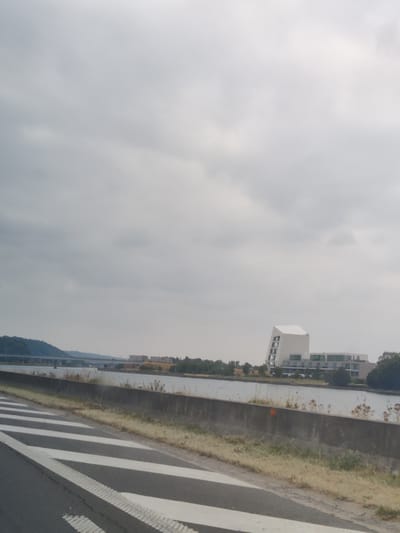 A grey, overcast sky dominates the upper portion of the image. In the distance, a body of water, likely a canal or river, is visible, with buildings and trees lining its banks. A prominent modern white building with an angular design stands out on the right side of the water. A bridge spans the water further in the background, connecting the two sides. The foreground of the image features a road with clear white lane markings and a concrete barrier. The scene appears to be a view from a road adjacent to a waterway in or near Hermalle-sous-Argenteau, Belgium.