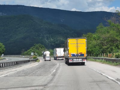 The image captures a scene on a highway near Vrachesh, Bulgaria, likely during daylight hours. The foreground shows a paved road with visible lane markings and some wear and tear. A bright yellow truck with "IVAN-OKIPROM" and "MEZHDUNARODEN TRANSPORT" written on its side is prominently featured in the right lane, moving away from the viewer. Behind it, in the same lane, is a white refrigerated trailer truck. The left lane shows a silver car driving ahead of a white tanker truck. The road curves to the left, with metal guardrails lining the median and the right side of the road. Beyond the road, a lush, tree-covered mountain range dominates the background. The sky is partly cloudy. The scene depicts traffic on a highway amidst a natural, mountainous landscape.