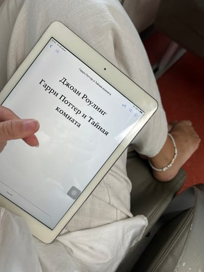 An overhead, medium shot captures a white iPad displaying a digital book. The book's title, "Гарри Поттер и Тайная комната" (Harry Potter and the Chamber of Secrets), is visible in large, bold Russian text, with the author's name, "Джоан Роулинг" (J.K. Rowling), above it. The timestamp in the top left corner of the iPad reads "16:03 Чт 25 июня". A person's left hand, with manicured fingernails, is positioned in the lower left quadrant of the image, appearing to hold the device. The iPad rests on a light-colored, textured fabric, likely clothing. In the background, to the right of the iPad, a person's bare foot is visible, adorned with a beaded anklet. The foot rests on a dark, possibly leather, surface, suggesting a seating area. The lighting is bright, indicating daylight.