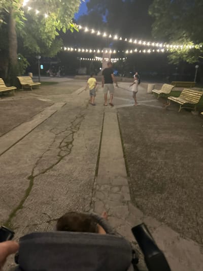 The image depicts an outdoor scene in the city of Parvomay, Bulgaria, at dusk. The foreground features a person's hands gripping what appears to be a stroller or a similar apparatus, looking down at the ground. The midground reveals a cracked concrete pathway with patches of greenery, leading towards a park-like area. In the park, two children are visible: a boy in a yellow shirt and striped shorts, and a girl in a white dress. An adult male in a black t-shirt and shorts is also present, seemingly accompanying the children. Several wooden park benches are scattered throughout the area, some appearing empty and others occupied by unseen individuals. Overhead, strings of lights illuminate the pathway, suggesting a festive or recreational atmosphere. The background shows trees and foliage, with the sky indicating the transition from day to night.