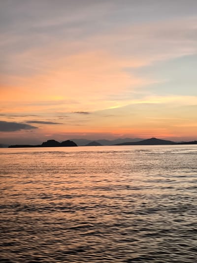 This is a vertical, eye-level, full shot of a serene seascape at sunset in Rinca Village, Indonesia. The sky is a gradient of soft oranges, pinks, and pale blues, with wispy clouds scattered across it. The horizon is dominated by the silhouettes of several islands and mountains, their shapes softened by the dusk. On the left, a prominent, jagged island stands out against the colorful sky. Further to the right, a conical mountain with a small structure at its peak is visible. The sea in the foreground is calm, reflecting the warm hues of the sky with gentle ripples. The overall mood is peaceful and tranquil, evoking the beauty of a tropical sunset. No people, objects, or text are visible in the image.
