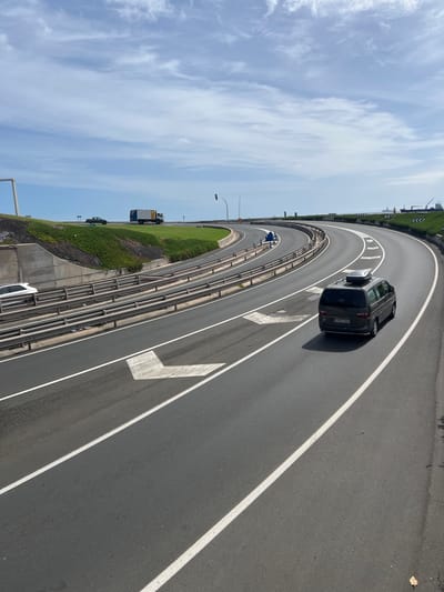 The image shows a section of highway in Las Palmas, Spain. A dark-colored vehicle is positioned on the left-hand side of the roadway, partially occupying the leftmost lane. The road has white lane markings and arrows indicating the direction of travel. A metal guardrail separates the highway from a grassy embankment. In the background, there are light-colored clouds against a blue sky. There are other vehicles on the road, including a truck and another car. The environment appears to be a bright, sunny day.