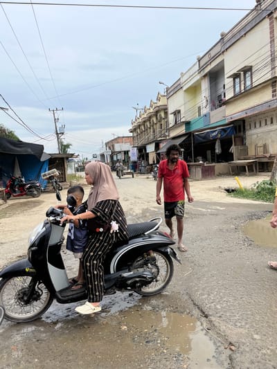 In Kota Kuala Simpang, Indonesia, a woman wearing a beige hijab and a black and white striped outfit is seated on a black motorcycle. She is holding onto the handlebars, and a young boy wearing a black and white jersey is positioned in front of her, also holding onto the handlebars. The motorcycle is parked on a wet, unpaved road with several large puddles. To the right of the motorcycle, a man with dark, curly hair and a red polo shirt is walking away from the camera, wearing black shorts and flip-flops. He appears to be gesturing with his right hand. In the background, there are several buildings with light-colored facades and ornate architectural details. Some of the buildings have awnings and open storefronts. Overhead, electrical wires crisscross the sky, supported by utility poles. A red scooter is parked to the left, partially obscured by a blue tarp. The overall scene depicts a street in a town with a mix of residential and commercial structures, under an overcast sky.