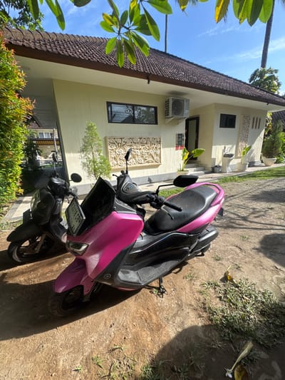 Two scooters, one black and one pink, are parked on a dirt and grass area in front of a light yellow building with a tiled roof. The building has several windows, an air conditioning unit, and decorative carvings on the exterior walls. In the background, trees and palm trees are visible under a bright blue sky with scattered clouds. The scooters are positioned close together, with the pink scooter in the foreground slightly obscuring the black one. The scene appears to be outdoors, possibly in a residential or tourist area, given the architecture and vegetation. The specific location in Karangasem, Indonesia, cannot be definitively determined from the image alone, but the visual cues suggest a tropical or subtropical environment. There are no people visible in the immediate vicinity of the scooters.