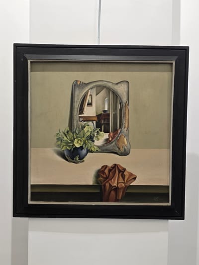 A framed painting is displayed against a white wall. The painting depicts a still life with a dark blue vase containing a green plant and a single yellow flower. To the right of the vase, a crumpled brown cloth rests on a beige surface. In the background, a peculiar mirror with a distressed, gold-trimmed frame reflects a room interior with furniture and a window. The painting is signed in the lower right corner with initials and a date, "D.B. '85". The artwork is hanging from a wire attached to the ceiling, and the bottom of the frame is visible against a white partition. The lighting suggests an indoor exhibition space. The city of Lom, Bulgaria, is not depicted in the image.