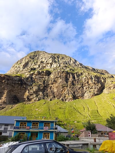 The image shows a mountainous landscape with a village in the foreground. A large, rocky mountain dominates the background, with green grassy slopes leading up to its base. Several buildings, including a blue guesthouse with "SHEPAN HOME STAY" signs, are clustered together in the village. Prayer flags are strung between some of the buildings. The sky is partly cloudy with blue patches. A black car is parked in the lower left corner of the image. The scene appears to be in a rural, mountainous region.